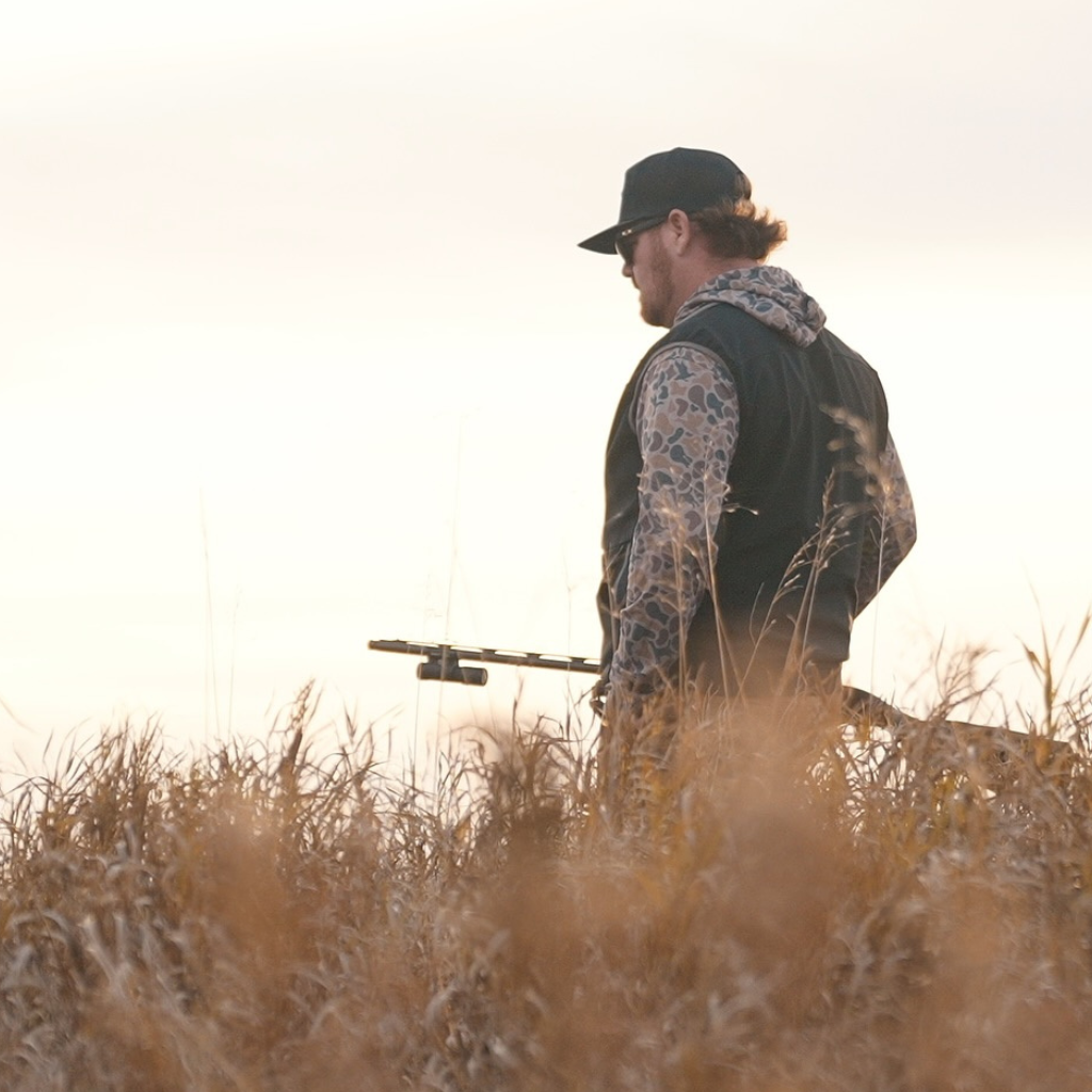 Man in camouflage clothing holding a rifle in a field at sunset