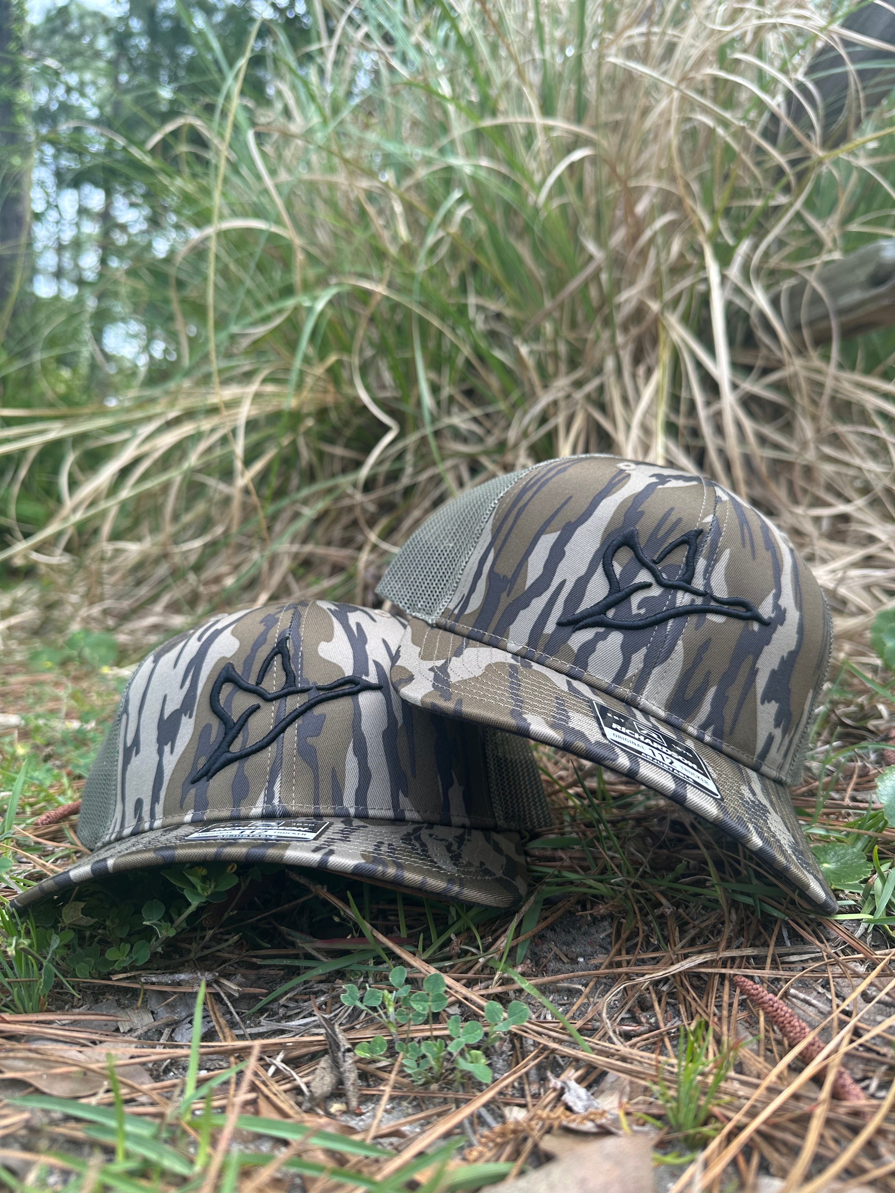 Two Richardson 112 Bottomland camo trucker hats with black 3D puff duck logo, mesh back, on grass.