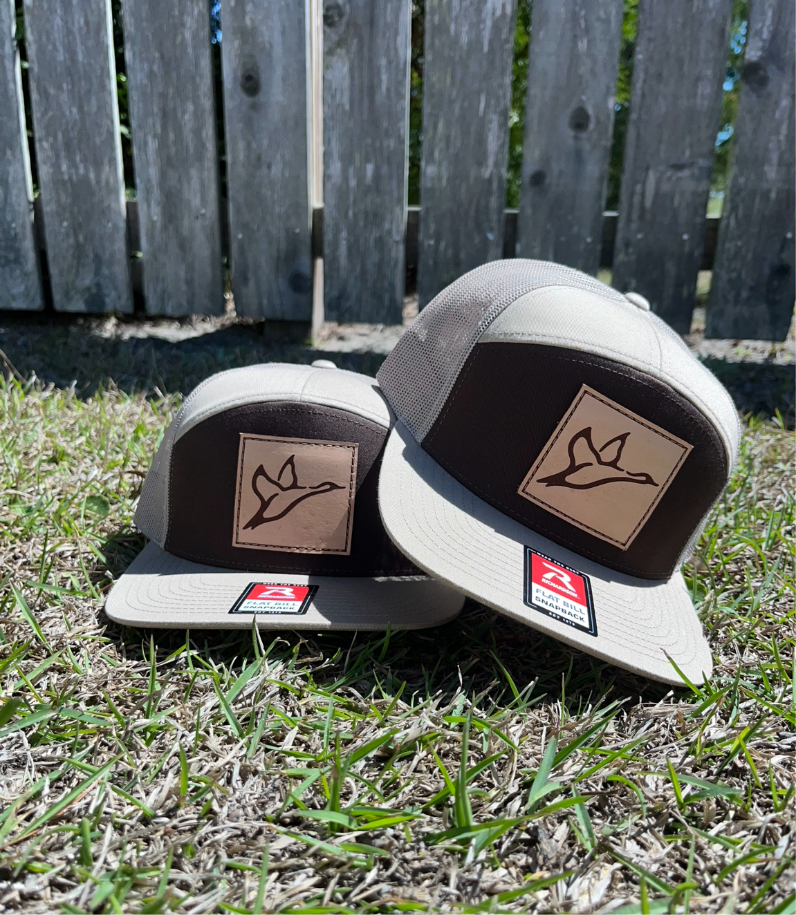 Two khaki and coffee 7-panel trucker hats with a duck icon on a leather patch, lying on grass.