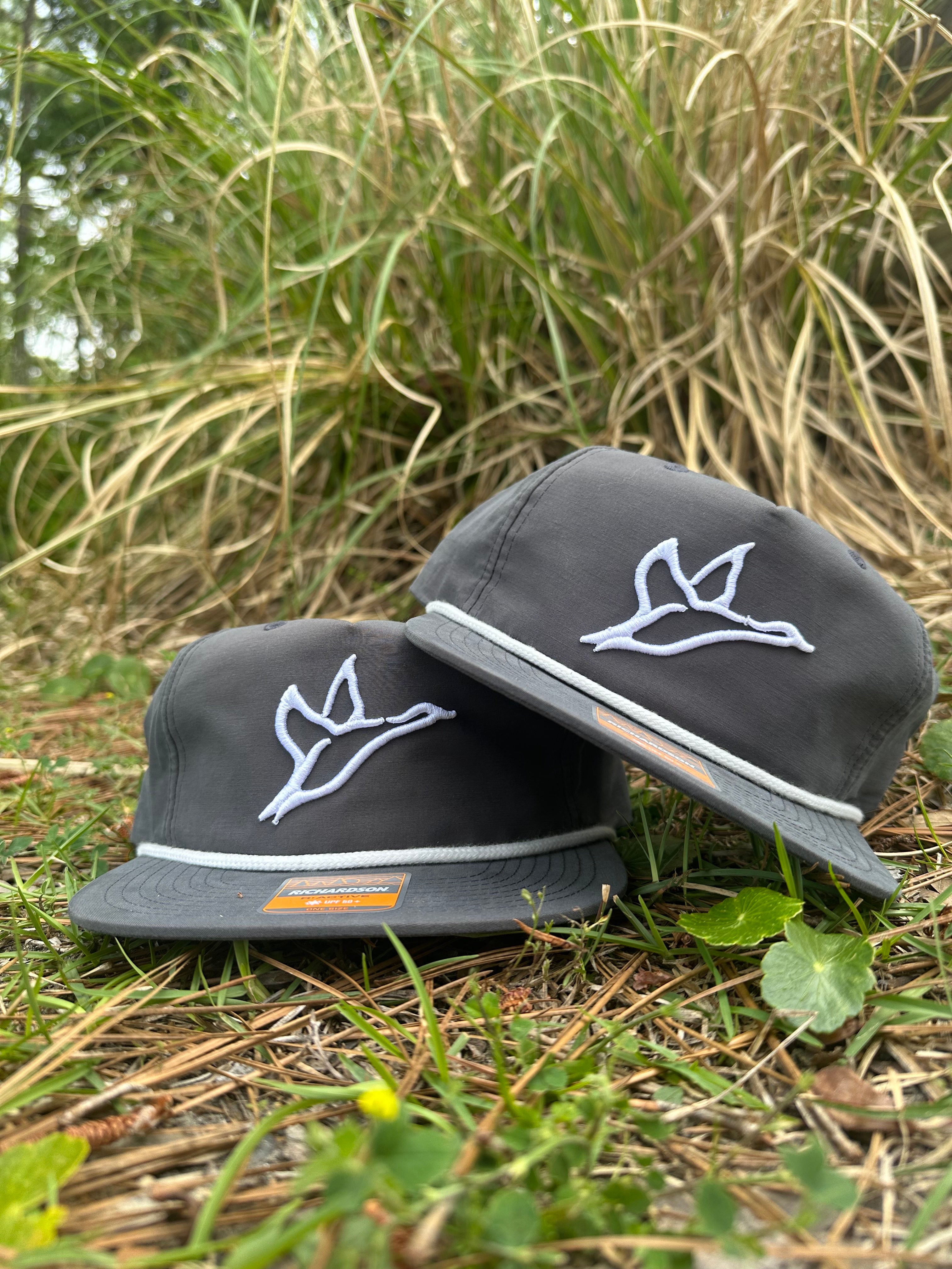 Two grey grandpa rope hats with white Combat Waterfowl duck icons in puff, resting on grass.