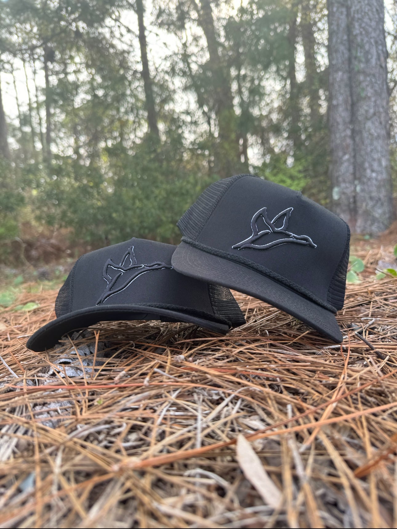 Two black Rope Hat - Black Ghost snapbacks with a subtle embroidered duck logo on pine needles.