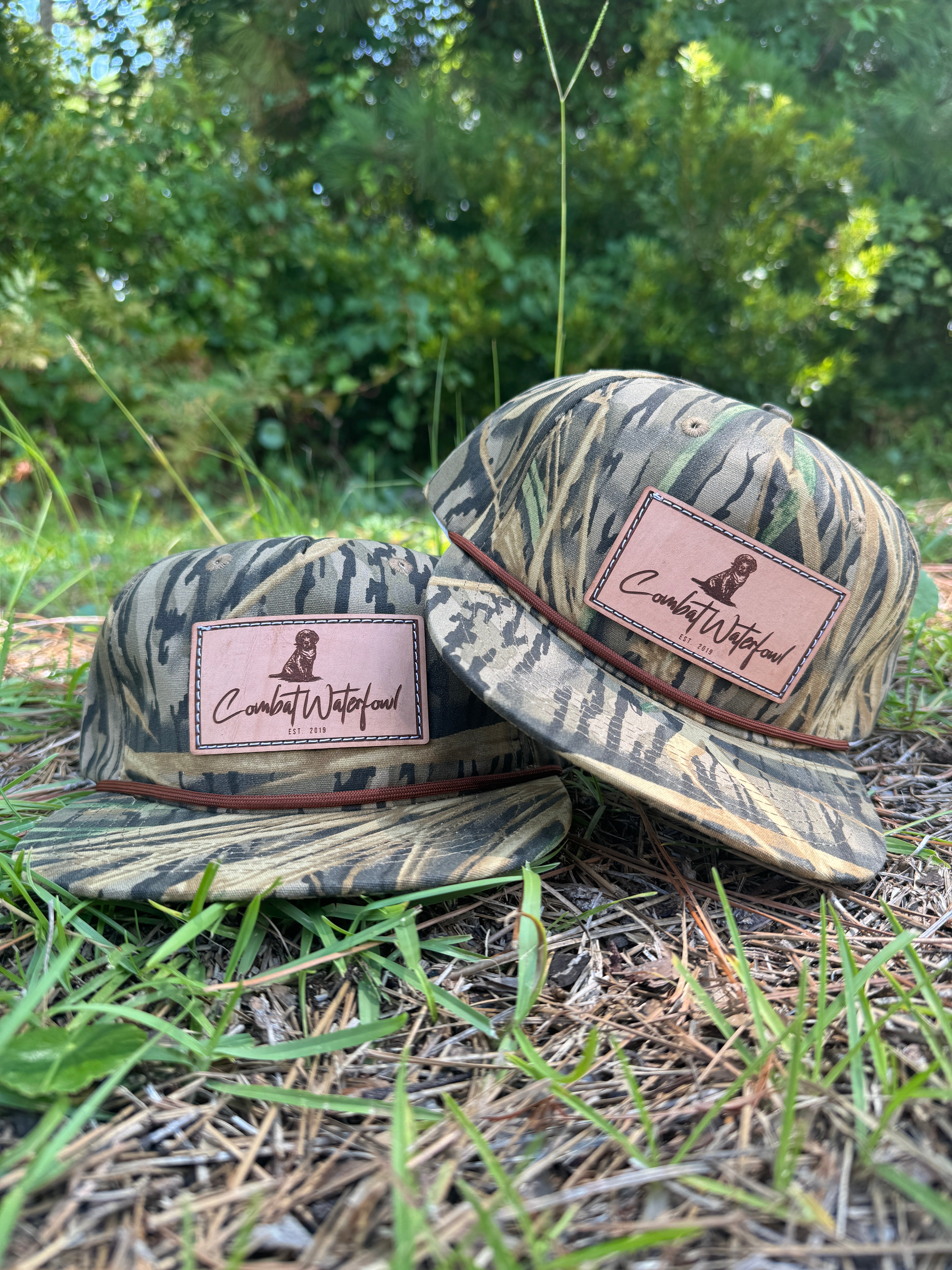 Two Goat Rope™ Mossy Oak™ Shadow Grass camo hats with genuine leather Lab patches, resting on grass.