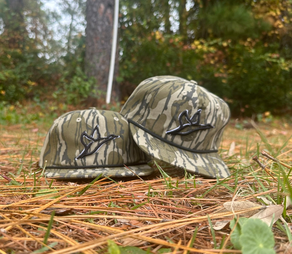 Two Goat Rope™ Mossy Oak™ Bottomland Ghost hats with duck emblems, one adult and one youth, on pine needles.