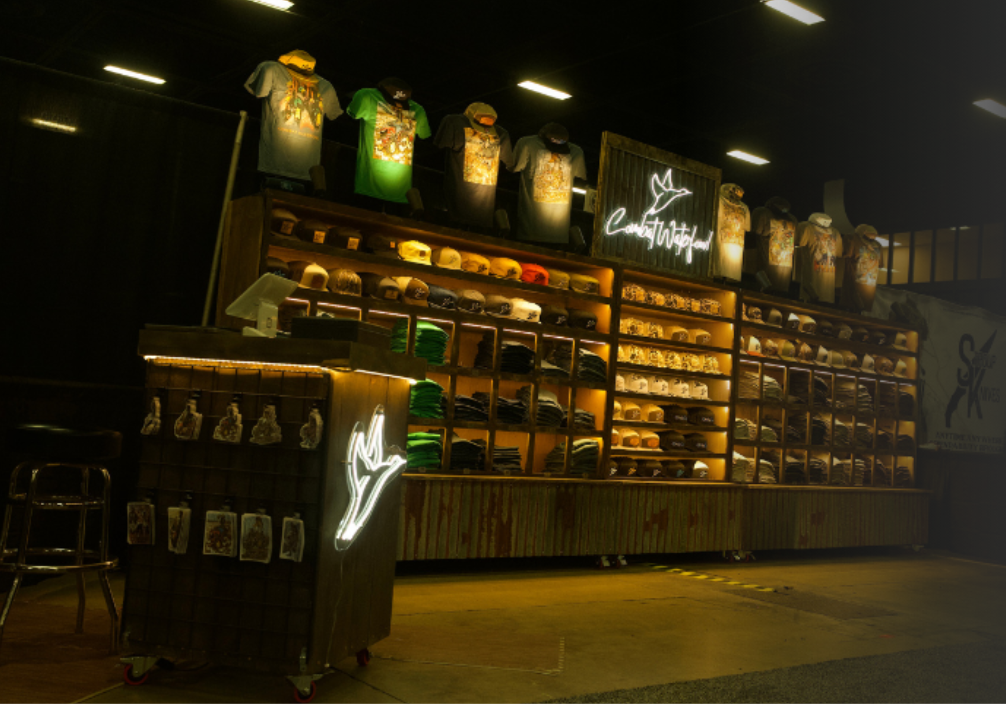 A retail display stand filled with apparel and accessories under warm lighting, featuring illuminated shirts, hats, and bags, with a dark background and a visible script logo on the display.