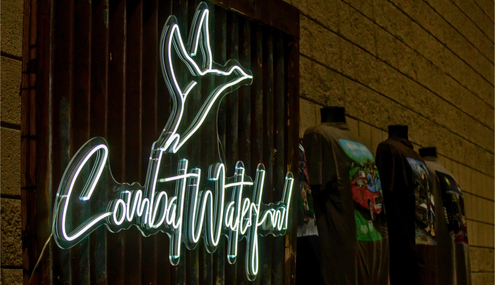 A glowing neon sign reading 'Combat Waterfowl' with a bird silhouette above it, mounted on a dark wooden door next to a tan brick wall and several mannequins wearing graphic T-shirts.